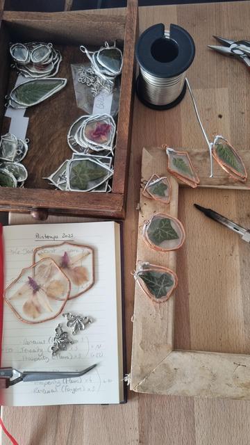English A worktop with glass pendants surrounded by a silver band like stained-glass, containing presses leaves and flowers. There are many more in a wooden drawer. Others are in the process of being finished, with the silver only covering the bail and the rest still in copper. The tin coil and soldering iron are visible.
•
Français Un plan de travail avec des pendentif en verre entourés d’une bande argentée comme des vitraux, contenant des feuilles et fleurs séchées. Il y en a quantité dans un tiroir en bois. D’autres sont en cours de finalisation, la partie argentée ne recouvre que l’attache et le reste est encore en cuivre. On aperçoit la bobine d’étain et le fer à souder.