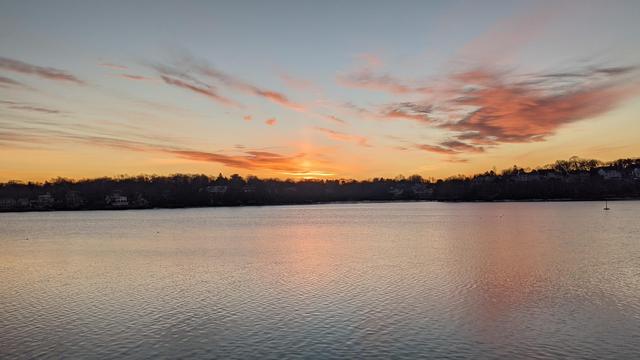 Historical Documentation in the form of a photograph of a rising sun over the ocean bay at the Historic Pioneer Village in Forest River Park, Salem, MA. The sky is filled with vibrant orange and pink hues, with the sun rising in the background. The water is calm and reflects the brilliant colors of the sky, creating a mirror-like effect. On the right side of the image, there is a small island with houses and trees on the shore. The island is surrounded by trees, and the water is still and calm. The overall mood of the photograph is peaceful and serene.