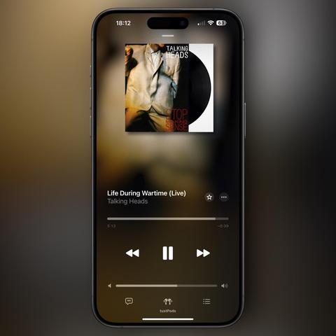 A smartphone screen displaying the album cover of "Stop Making Sense" by Talking Heads, specifically the song "Life During Wartime (Live)." The playback time shows 5:13, accompanied by playback controls. The background is a blurred gradient.