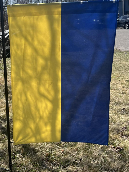 The blue-and-yellow Ukraine flag hangs vertically from a banner holder in our front yard, where the grass has not yet turned green in the spring.
