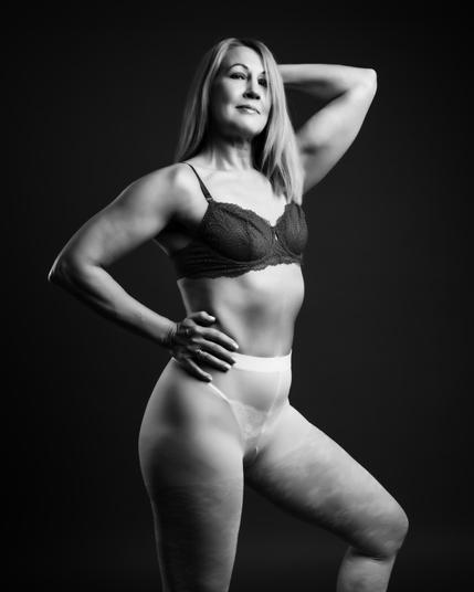 A black and white studio portrait of a confident and fit +40 female model wearing lace lingerie and sheer tights, posing gracefully to showcase strength and elegance.