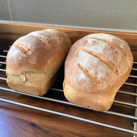2 loaves of bread on a cooling rack, set on a dark wooden counter.