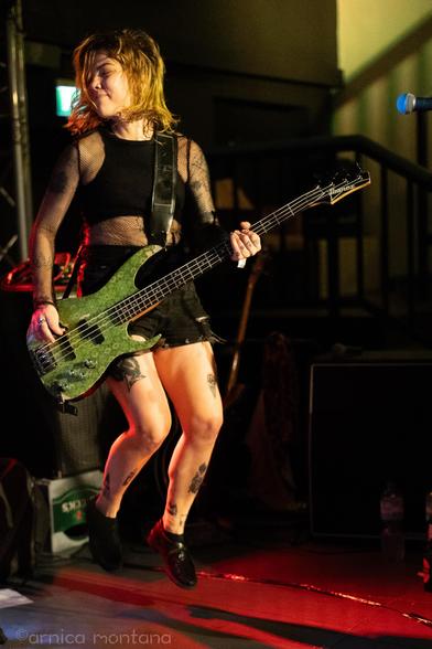 A musician performing on stage, playing a green bass guitar while jumping, wearing a fishnet top and a skirt. Tattoos are visible on her legs, and the stage is dimly lit with various background elements.