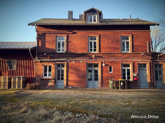 Ehemaliger Bahnhof von Laubach in Hessen, ein historisches Backsteingebäude mit blauen Türen und Fenstern, im warmen Licht der tief stehenden Sonne. Im Vordergrund stehen Mülltonnen, Holzpaletten und ein leicht frostiger Boden, während lange Schatten die Fassade betonen. Die Szene wirkt nostalgisch und erinnert an die vergangene Zeit des Bahnverkehrs.