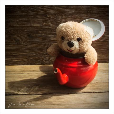 The image features an adorable teddy bear peeking out from inside a vibrant red teapot. The bear's soft fur and friendly expression exude warmth and playfulness. The teapot, with its shiny surface and gracefully curved spout, adds a whimsical touch to the scene, creating a delightful contrast between the functional kitchen object and the cuddly toy.
The background of weathered wood enhances the rustic charm of the composition, while casting gentle shadows that suggest a play of light and time. This setup evokes a sense of nostalgia, potentially conjuring childhood memories or a feeling of comfort. The overall arrangement sparks curiosity—blurring the lines between reality and imagination, as the teapot serves not in its intended capacity but rather as a cozy spot for the teddy bear.

