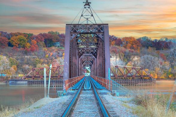 A railroad track leads to a truss bridge crossing the Mississippi River. Set against a backdrop of vibrant autumn foliage.