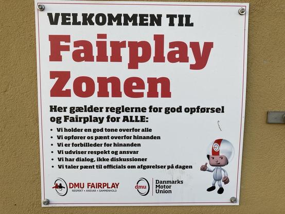 Skilt på en væg med teksten 'VELKOMMEN TIL Fairplay Zonen'. Skiltet beskriver regler for god opførsel og fairplay, herunder at vise respekt, være gode forbilleder og tale pænt. Nederst på skiltet ses logoet for Danmarks Motor Union (DMU) samt en tegneseriefigur iført hvid køredragt og hjelm, der giver en thumbs-up
