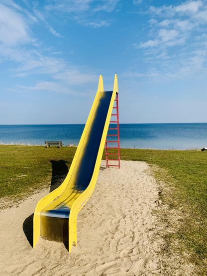 Eine gelbe Spielplatzrutsche mit roter Leiter, im Hintergrund das Meer und ein blauer Himmel.