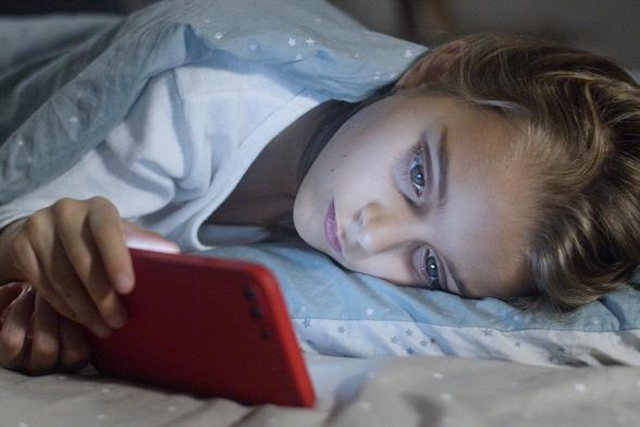 A person is lying in bed under a light blue blanket patterned with white stars. The person’s head rests partially visible on a pillow, while their hand holds a red smartphone. The dim, soft lighting suggests a relaxed atmosphere, with the individual engaging with their phone, possibly before sleeping or after waking.