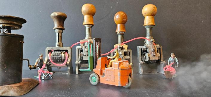 Oude stempels, van El Casco, een oude metalen miniatuur seinbel van een oude speelgoedtreinset en wat tinnen miniatuurfiguurtjes, die met een metalen vorkheftruck druk in de weer zijn 