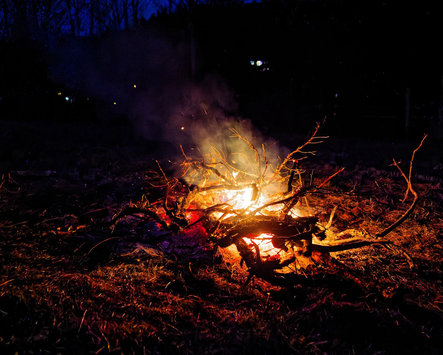 A fire with a bright central region with a large stick running through it with branches going out in different directions and a plume of illuminated smoke. Lights on houses across the creek are visible in a dark expanse above the fire with a patch of dark blue sky visible through the trees to the upper left. A fire with a bright central region with a large stick running through it with branches going out in different directions and a plume of illuminated smoke. Lights on houses across the creek are visible in a dark expanse above the fire with a patch of dark blue sky visible through the trees to the upper left.