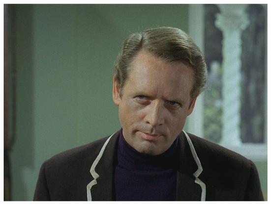 Still image of the character known as Number Six, played by Patrick McGoohan, in the TV series "The Prisoner".
Pictured from the chest up, he wears a black blazer with white piping over a black shirt. He looks camera left with an expression suggesting suspicion.