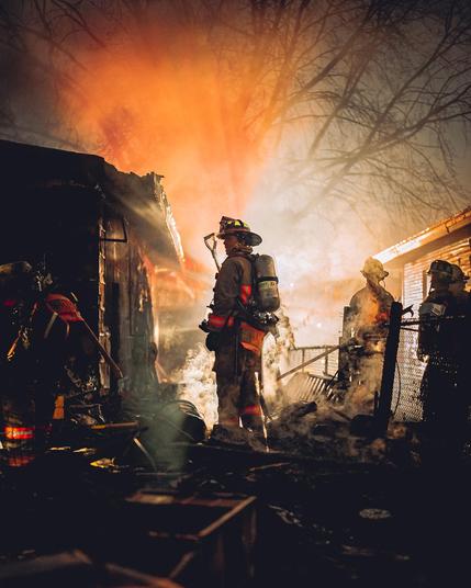 A dramatic nighttime photograph of firefighters battling a house fire. A firefighter wearing full protective gear, including a helmet and oxygen tank, stands in the center of the frame surrounded by smoke and glowing embers. The remains of a burning structure are visible on the left, with charred debris scattered around. Other firefighters work in the background, silhouetted against the intense orange and yellow glow of the flames. Bare tree branches stretch overhead, adding to the dramatic atmosphere.