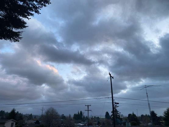 Photo of a stormy sky with blue hues popping in from behind the clouds. A telephone pole and wire stand in the foreground with the tops of houses and a town in the midground. 