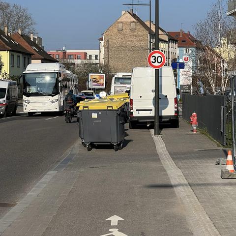 Une camionnette blanche stationnée à cheval sur le trottoir et la piste cyclable, tandis que le reste de la piste cyclable est occupé par des poubelles jaunes. Un vélo est obligé de se mettre sur la chaussée pour se déplacer.