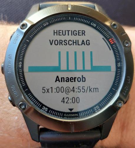 Man sieht eine Garmin fenix Uhr, auf der der heutige Trainingsvorschlag angezeigt wird. Oben steht der Text "Heutiger Vorschlag", darunter sieht man eine Grafik, die den Tempoverlauf des Trainings verdeutlichen soll und darunter steht als Text "Anaerob", "5x1.00@4:55/km" und "42:00".