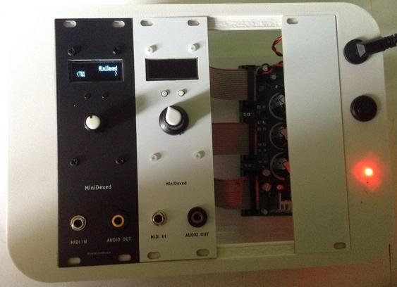 Photo of two 8HP DIY EuroRack modules, both essentially the same design, but one black, one white.  Both with a small OLED display at the top, encoder in the middle, and two TRS sockets at the bottom.  The black one on the left is powered up showing the "MiniDexed - TG1" display.  Both modules are in my cake-tub-EuroRack box for testing.
