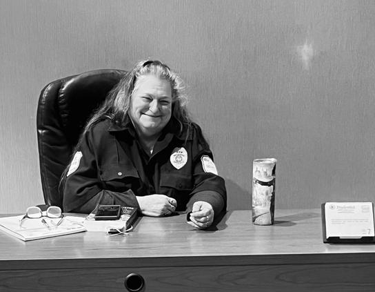 A black and white portrait of a woman security guard sitting at a desk with her glasses, a book, travel coffee mug, and desk calendar. She has a wonderful smile. 