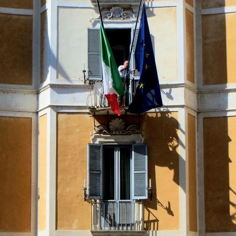 A man is standing on a small balcony to untangle an EU Flag attached to the building facade.