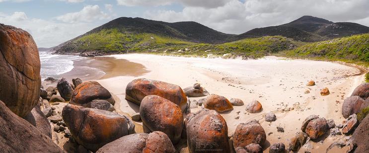 A panoramic view of Whiskey Bay, VIC Australia, featuring large, rust-coloured boulders scattered across a sandy shoreline. The beach curves gently toward the ocean, with waves lapping at the shore, and is framed by lush green hills and a partly cloudy sky, creating a serene coastal scene.