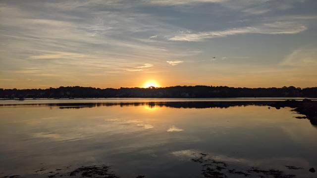 Historical Documentation in the form of a photograph of a rising sun over the ocean bay at the Historic Pioneer Village in Forest River Park, Salem, MA. The sky is filled with orange and yellow hues, with the sun peeking over the hills on the horizon. The water is calm and still, reflecting the colors of the sky. On the right side of the image, there is a rocky shoreline with trees and buildings visible in the distance. The sun is setting, casting a warm glow over the scene. The overall mood of the photograph is peaceful and serene.