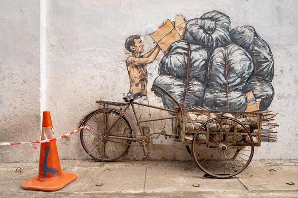 A photograph of a wall mural in Ipoh, Malaysia, where art and reality merge. The mural depicts a man straining to balance an overloaded tricycle stacked high with goods, while a real tricycle is positioned in front of the painting, seamlessly blending two and three dimensions. In the foreground, an orange road cone stands alone, adding a modern contrast to the scene.