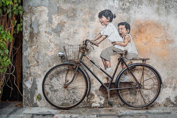 A photograph of a well-known street art mural in George Town, Penang, Malaysia, depicting two children riding a bicycle. Their painted figures appear full of life, with joyful expressions and a sense of movement, blending naturally with the real, mounted bicycle attached to the wall. Created by Lithuanian artist Ernest Zacharevic, this mural has become an iconic feature of George Town’s cultural landscape.