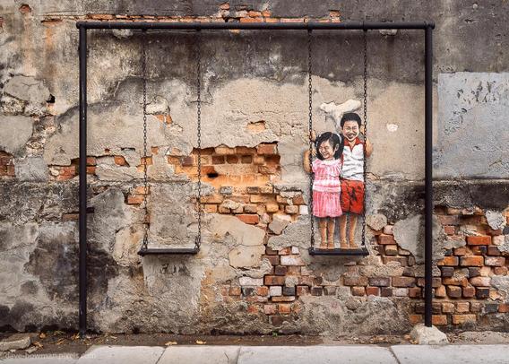 A photograph of street art in the backstreets of George Town, Penang, Malaysia, showing two children smiling and playing on a swing. The painted figures of a young boy and girl appear to be sitting on a real swing attached to the weathered wall, blending the art with the physical surroundings. The scene feels playful and nostalgic, bringing a sense of life and movement to the otherwise aged and textured wall.