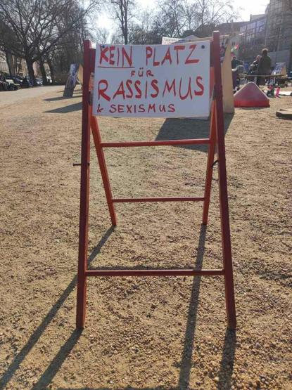 Ein Stuhl ohne Sitzfläche an dem ein Schild befestigt ist, auf dem "Kein Platz für Rassismus & Sexismus" steht. Im Hintergrund ist das Protestcamp am Oranienplatz sichtbar.