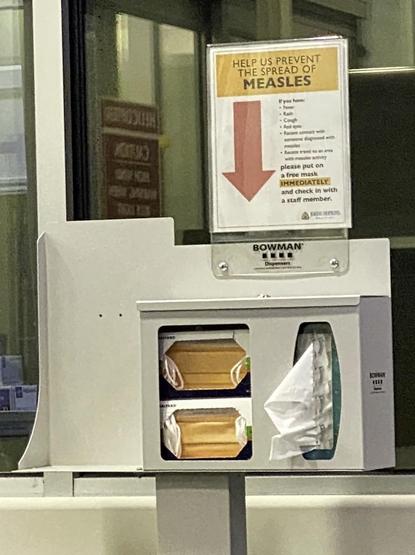 Sign text: "Help us prevent the spread of MEASLES. If you have: fever, rash, cough, red eyes, recent contacts with someone diagnosed with measles, recent travel to an area with measles activity, please put on a free mask immediately and check in with a staff member" on a box with masks and tissues. 