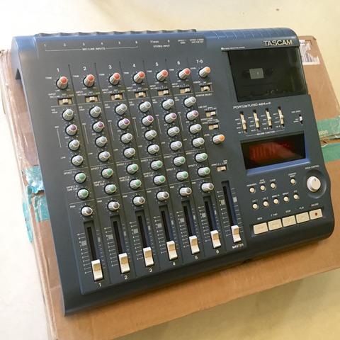 A very clean Tascam 424mkIII multitrack cassette recorder sits atop a brown, pre-used cardboard shipping box on a white linoleum floor.