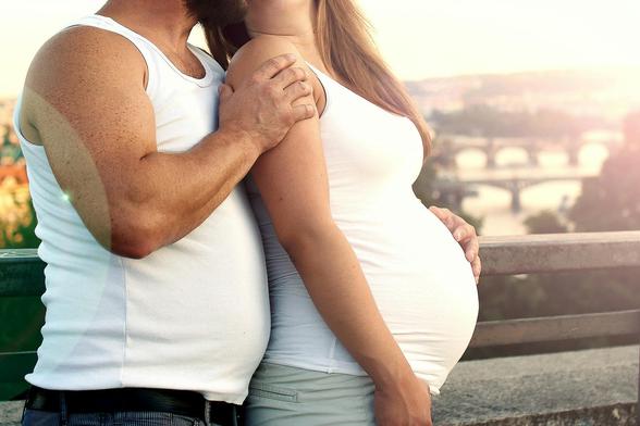A pregnant woman and a man standing close together outdoors. Both are wearing white sleeveless tops. The man has his arm around the woman's shoulder, and the woman is holding her belly. In the background, there is a scenic view of a river with several bridges.