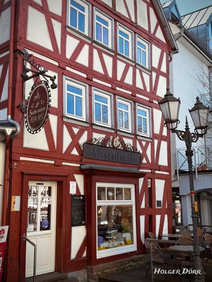 Ein charmantes Fachwerkhaus mit rot-weißer Fassade beherbergt das „Altstadtcafé“ in Laubach, Hessen. Über dem Eingang hängt ein nostalgisches Schild mit der Aufschrift „Zuckerbäcker Haas“. Die große Fensterfront zeigt dekorative Elemente und Backwaren. Eine handgeschriebene Tafel neben der Tür bewirbt „Sachertorte“ und „Granatsplitter“. Vor dem Café stehen Tische und Stühle auf einer kleinen Außenterrasse. Rechts im Bild befinden sich historische Straßenlaternen. Die Fenster spiegeln den blauen Himmel wider, was auf einen sonnigen Tag hinweist.