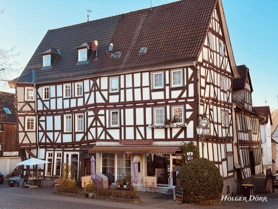 Ein traditionelles Fachwerkhaus in Laubach, Hessen, mit einer Bäckerei und einem Café im Erdgeschoss. Vor dem Eingang stehen Tische und Stühle mit geschlossenen Sonnenschirmen auf einer kleinen Terrasse. Ein Schild weist auf Kaffeespezialitäten hin. Die Fassade ist weiß mit dunklen Holzbalken, und das Gebäude hat mehrere Fenster mit weißen Rahmen. Die Sonne wirft warme Schatten auf das Kopfsteinpflaster.