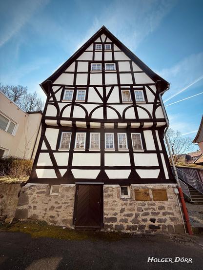 Historisches Fachwerkhaus in Laubach, Hessen, bekannt als das Strumpfweberhaus - das älteste Wohnhaus der Stadt, um 1450 errichtet. Das Gebäude hat eine markante Schiefstellung und besteht aus dunklen Holzbalken mit weiß verputzten Flächen. Der untere Teil ist aus Bruchsteinmauerwerk, während der obere Teil die typische Fachwerkarchitektur aufweist. Rechts führt eine Treppe entlang des Gebäudes, und an der Fassade befinden sich Informationstafeln. Der Himmel ist blau mit leichten Wolken und Kondensstreifen.