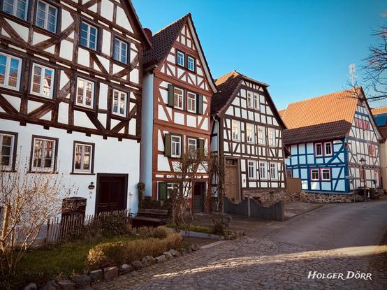 Eine malerische Fachwerkhauszeile in Laubach, Hessen, bei strahlend blauem Himmel. Die historischen Gebäude sind in verschiedenen Farbkombinationen gestaltet, darunter ein auffälliges rot-weißes Fachwerkhaus mit grünen Fensterläden. Sonnenlicht betont die Strukturen der Häuser und verleiht der Szene eine warme, einladende Atmosphäre. Im Vordergrund ein gepflasterter Eingangsbereich mit einer kleinen Bank und etwas Grün. Die Idylle spiegelt den traditionellen Charme der hessischen Fachwerkstädte wider.