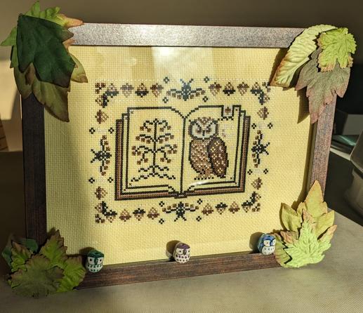 Completed cross stitch on a sunny yellow fabric. The stitches show a book surrounded by foresty elements. On the left side of the open book is a sparse tree and on the right is an owl.