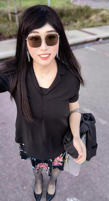 A woman with long dark hair takes a selfie outdoors in a parking lot. She is wearing stylish sunglasses, a black collared shirt, a floral-patterned skirt, black tights, and black heels. She has a smile on her face and is accessorized with dangling earrings and a delicate necklace. In her left hand, she carries a black jacket and a light-colored handbag. The background includes pavement, a curb, and some greenery.