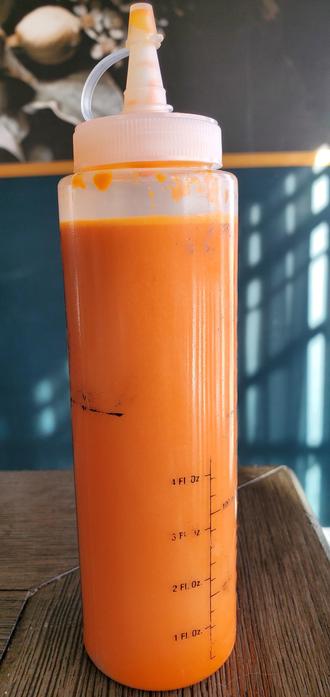 A plastic container filled with vibrant orange hot sauce in the foreground on a table with a green wall behind it.