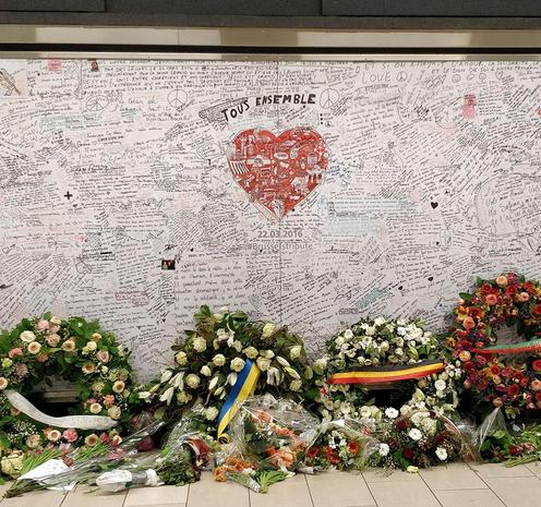 Mur recouvert de centaines de messages de solidarité, avec un grand cœur rouge au centre sur lequel est inscrit "Tous ensemble". Devant le mur, plusieurs couronnes de fleurs, bouquets et rubans, en hommage aux victimes de l’attentat du 22 mars 2016 à la station Maelbeek, Bruxelles.

