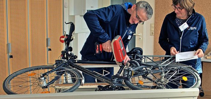 ADFC-Mitarbeiter*innen stehen vor einem auf dem Tisch liegenden Fahrrad. Die Frau hat ein Blatt mit dem Codierpass in der Hand, der Mann setzt das Codiergerät am Rahmen an, um es zu codieren.