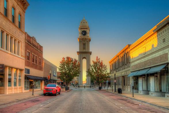 A clock tower stands prominently in the center of a quiet street, framed by two rows of quaint buildings reflecting the warm glow of the setting sun
