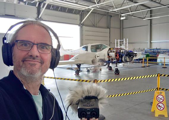 Rainer Suckow steht in einer Halle, in dem das Flugzeug Apus I-2 einen Testlauf absolviert. In der Hand hält er ein Aufnahmegerät.