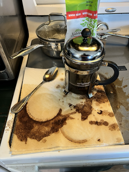 A coffee press on a stovetop with lots of spilled coffee, napkins and a spoon. 