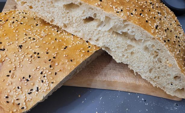 Farbfoto: Auf einem Schneidebrett liegt ein halbwegs rundes Fladenbrot aus hellem Mehl mit der typischen lockeren Krume, der goldgelben Kruste mit Sesam und Schwarzkümmel bestreut. 