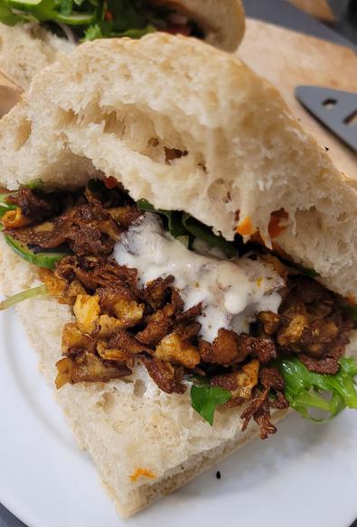 Farbfoto: Auf einem weißen Teller liegt ein Viertel vom Fladenbrot des ersten Tröts, gefüllt mit Salat (Blattsalat, Tomaten, Gurken, Petersilie, rote Zwiebeln sind nicht wirklich sichtbar...), dem marinierten und gebratenen Räuchertofu, etwas Joghurtsauce und irgendwo versteckt auch etwas Ajvar. Im Hintergrund liegt, nur angeschnitten sichtbar, ein zweites solches Sandwich, ein Pfannenwender und Küchenkram. 