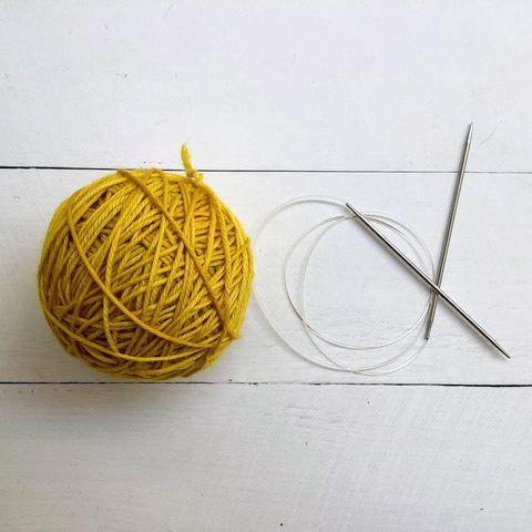 Flat lay view of a bright yellow ball of yarn next to a circular needle on a white plank background.