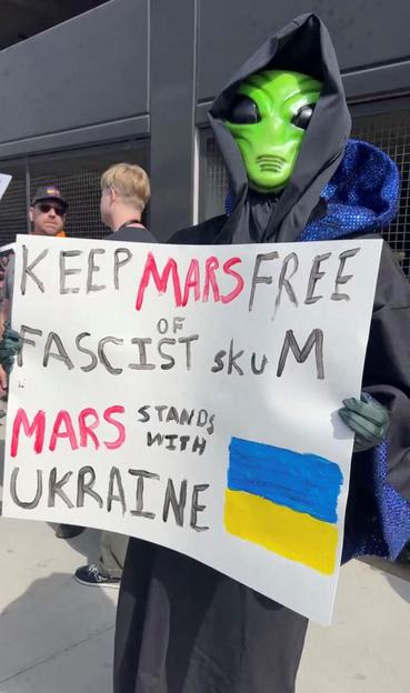 A person in a "green man" mask holds a sign reading:

"Keep Mars free of fascist skum, 

Mars stands with Ukraine"