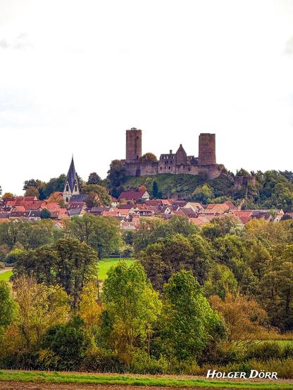 Die Burg Münzenberg thront auf einem Hügel über der gleichnamigen Stadt in der hessischen Wetterau. Im Vordergrund liegt ein ruhiges Städtchen mit rot gedeckten Fachwerkhäusern und einer auffälligen Kirche mit spitzem Turm. Die herbstlich gefärbten Bäume und das weiche Licht verleihen der Szene eine friedliche, fast zeitlose Stimmung.