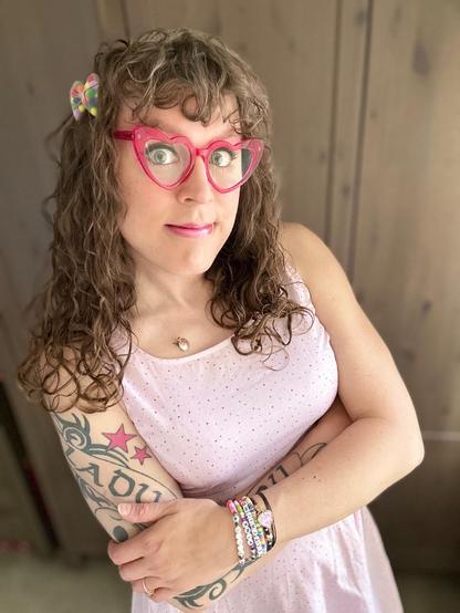 Me in a light pink dress with small multicolored polka dot stars, and pink heart shaped glasses. I have curly bangs and long brown curly hair, in which there are see-through mesh bows filled with small multicolored balls of fabric. I’m wearing multiple bright friendship bracelets, and my arms are crossed as I smile at the camera. I’m also wearing sparkly pink lipstick and dark eyeliner. And my right arm is covered in with my tattoo sleeve. I’m Cute af!